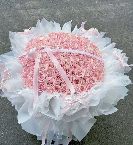 A large heart-shaped bouquet of pink roses wrapped in white tulle paper and tied with a pink ribbon, placed on a gray concrete surface.
