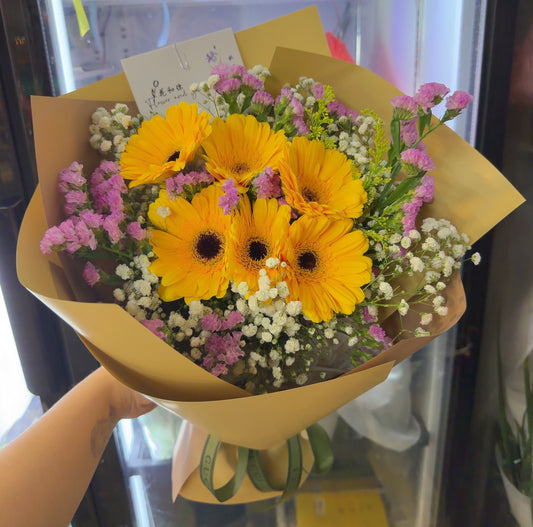 A hand holding a wrapped bouquet of yellow gerbera daisies, pink carnations, and white baby's breath flowers in brown paper wrapping, with a green ribbon at the bottom.