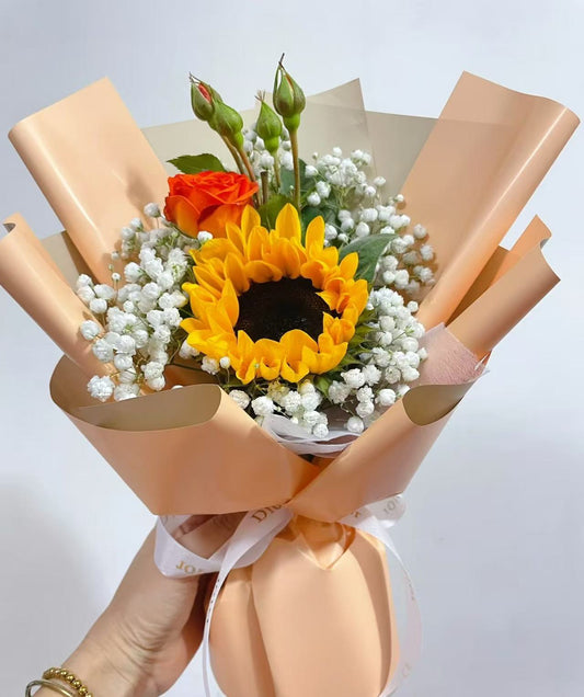 A hand-held flower bouquet featuring a large yellow sunflower, a red rose bud, baby's breath, and green stems, wrapped in peach-colored paper with a white ribbon.