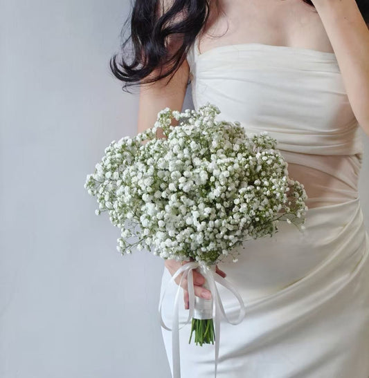 A person in a strapless ivory dress holding a large bouquet of white baby's breath flowers with a white ribbon, against a plain light gray background.