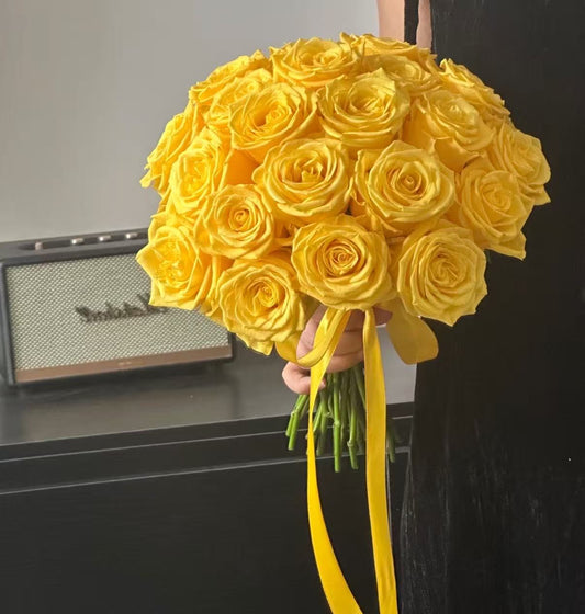 A large bouquet of bright yellow roses held by a person, with long yellow ribbon stems visible against an indoor background featuring a Marshall speaker.