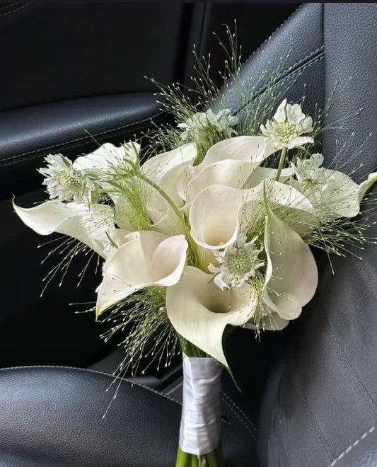 A bouquet of white calla lilies and green filler flowers wrapped in white ribbon, resting on a black leather car seat.