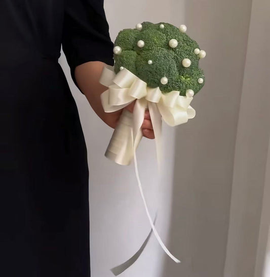 A person in a black dress holds a decorative bouquet made from a green broccoli floret wrapped with a large cream satin ribbon and embellished with white faux pearls, against a plain light gray background.