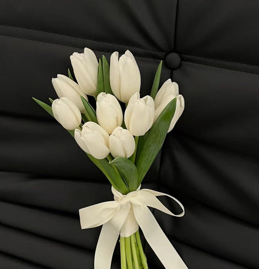 A bouquet of white tulips with green stems and leaves, tied with a white satin ribbon, set against a black quilted fabric background.