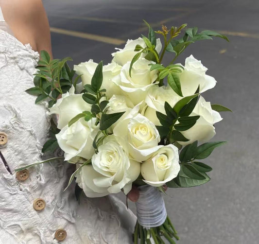 A bouquet of white roses with green leaves, held by a person wearing a textured white lace dress outdoors on a paved surface.