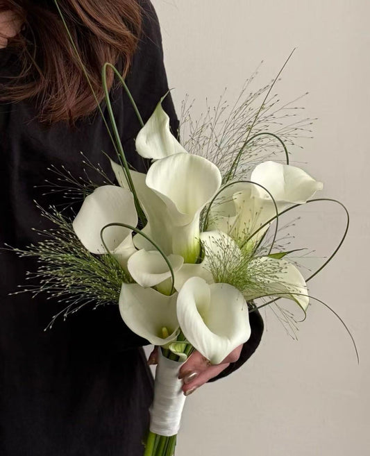 A bouquet of white calla lilies and delicate green filler grasses held by a person wearing a black top against a plain light beige wall.