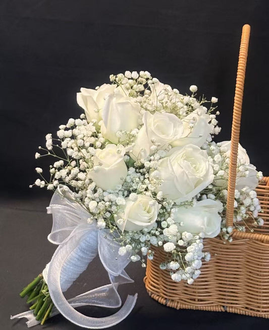 A bouquet of white roses and baby's breath tied with a sheer white ribbon, displayed next to a wicker basket against a dark background.