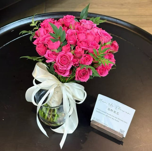 A vibrant bouquet of pink roses in a clear glass vase with a white ribbon bow, placed on a dark round tray next to a small printed card.