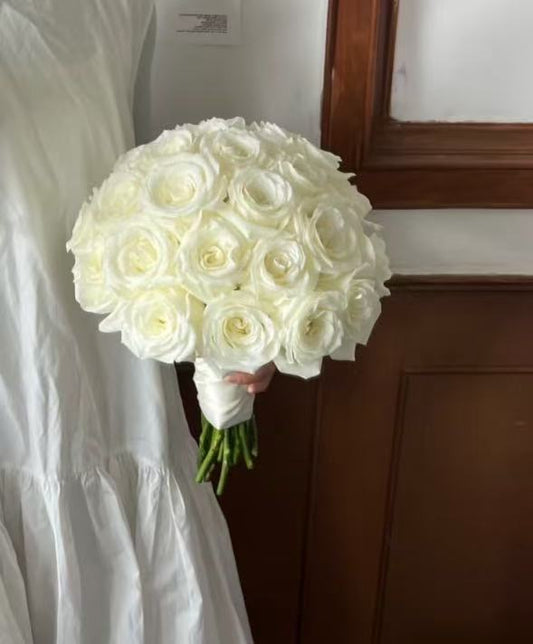 A large round bouquet of white roses held by a person wearing a white dress, standing indoors next to a dark wood door.