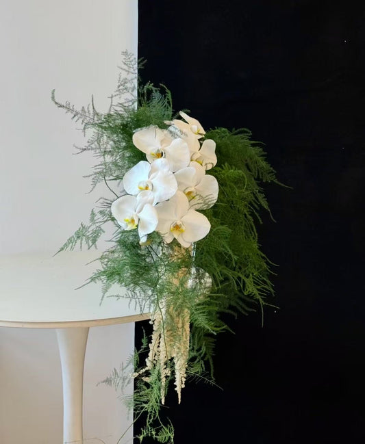 A cascading floral arrangement featuring white orchids, green ferns, and delicate cream seed pods, draped over a white table against a split white and black background.