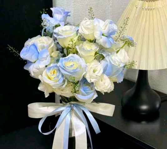 A bouquet of artificial light blue and white roses tied with a pale blue ribbon, displayed on a dark table next to a black lamp.