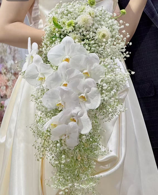 A bridal bouquet featuring large white orchids and delicate white baby's breath, held by a bride in a cream satin wedding dress next to a dark navy suit.