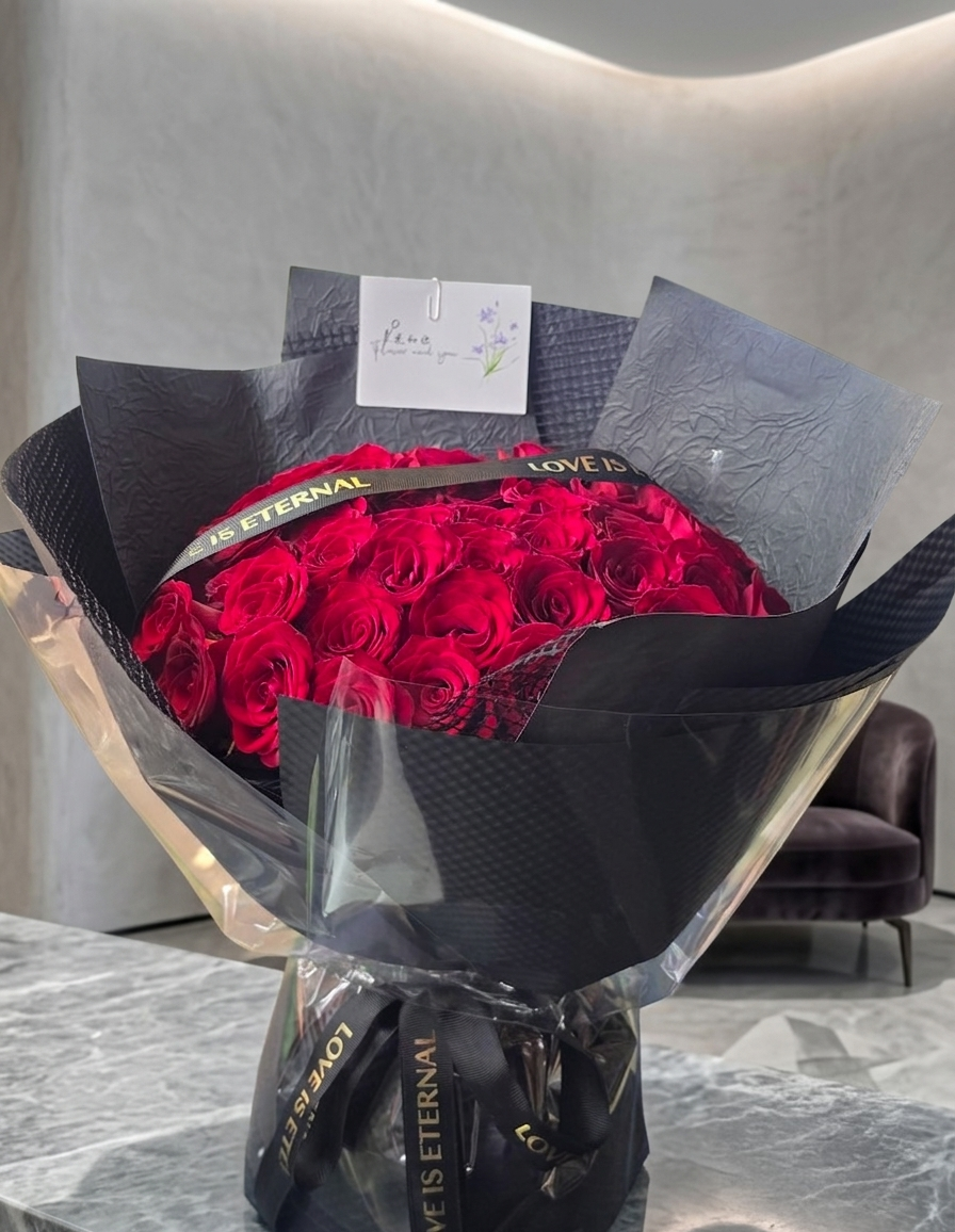 A large round bouquet of deep red roses wrapped in black tissue paper and clear cellophane, with a black ribbon printed 'LOVE IS ETERNAL' and a small card reading 'Me: I love you with all my heart'.