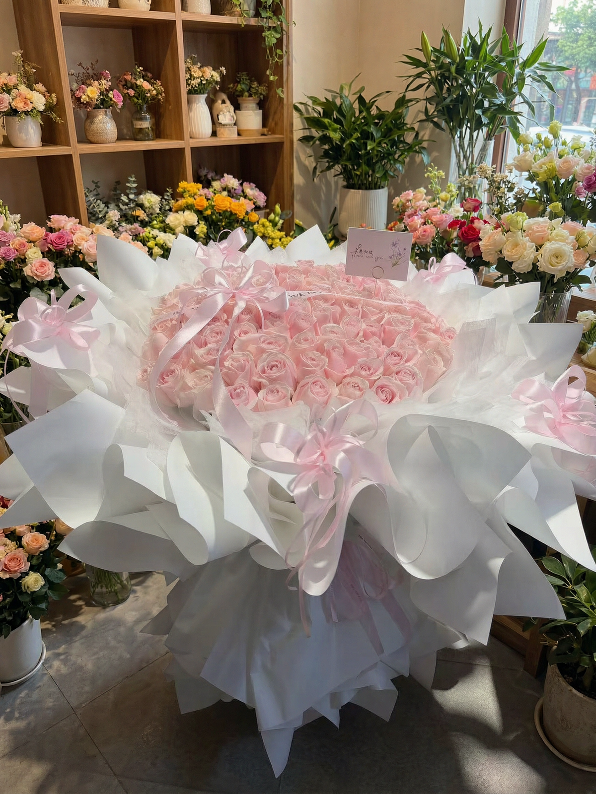 A large pink rose bouquet wrapped in white paper and tied with pink ribbons, displayed in a flower shop with shelves of flowers in the background.