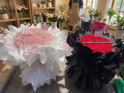 Two large flower bouquets displayed on a table in a flower shop. The bouquet on the left is made of pink roses wrapped in white paper with pink ribbon, and the bouquet on the right is made of red roses wrapped in black paper with black ribbon.