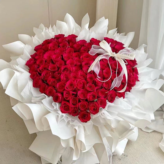 A heart-shaped bouquet of red roses wrapped in white tissue paper and tied with a white ribbon, photographed indoors against a light neutral background.