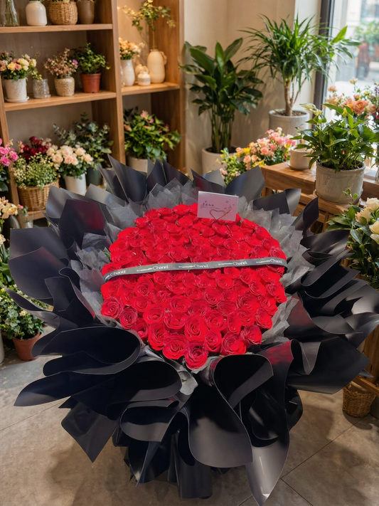 A large heart-shaped floral arrangement made of red roses, wrapped in black paper and a silver band, displayed in a flower shop with shelves of flowers and potted plants in the background.