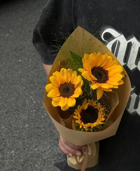 A bouquet of three yellow sunflowers wrapped in brown kraft paper, held by a person wearing a black graphic t-shirt against a gray asphalt background.