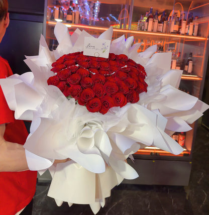 A large bouquet of red roses wrapped in white paper, held by a person wearing a red shirt in a retail or gift shop setting with illuminated shelves in the background.