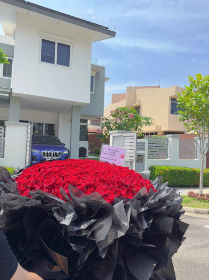A large bouquet of red roses wrapped in black paper, photographed outdoors in front of a modern house and gate. A small sign is visible among the flowers, and part of a person's arm appears at the lower left edge.