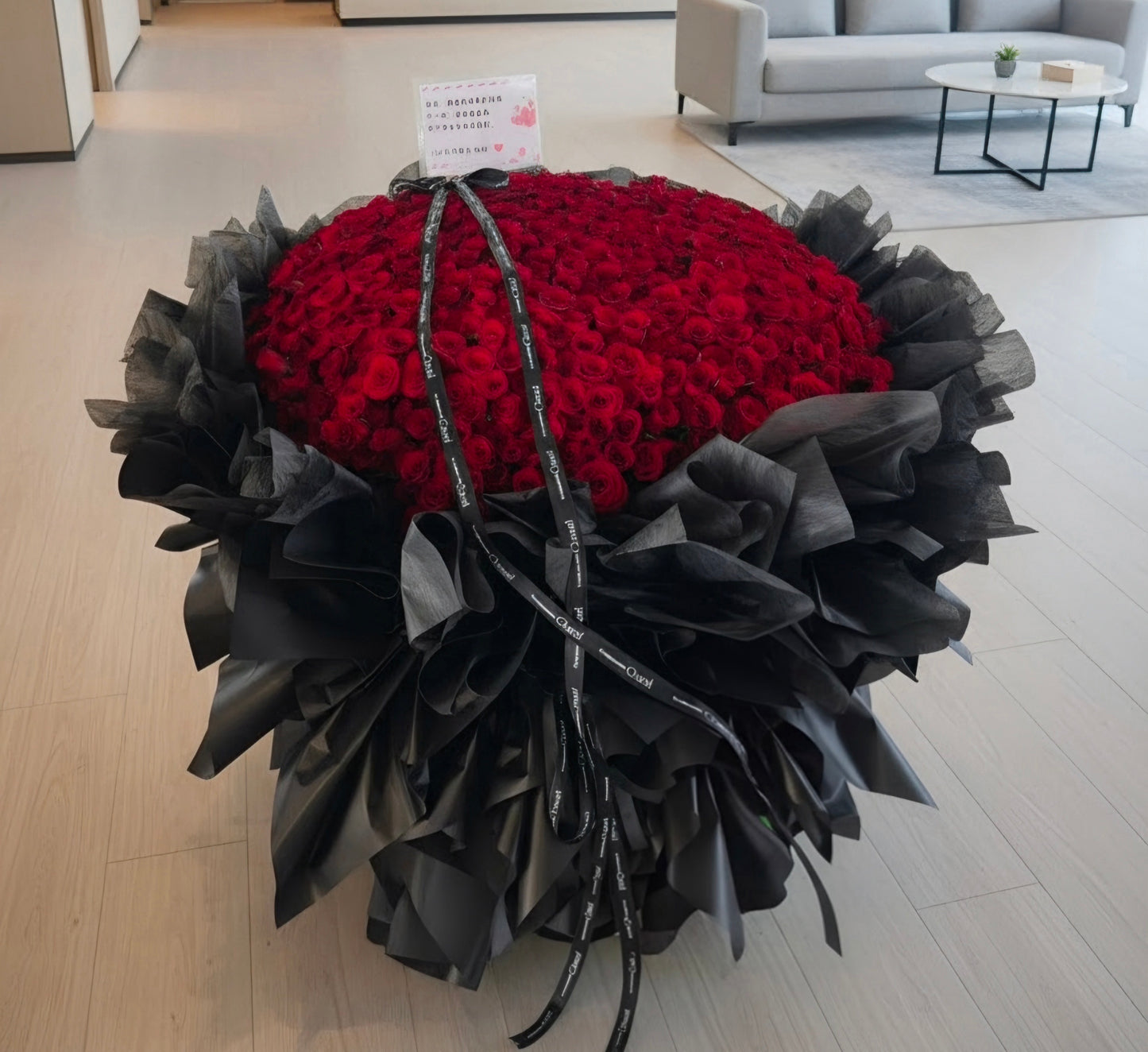 A large heart-shaped flower arrangement made of red roses, wrapped in black paper and tied with black ribbons, placed in a modern living room setting.