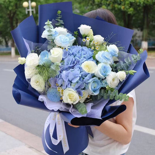 A person holding a large bouquet of light blue and white flowers wrapped in dark blue paper, photographed outdoors on a street with trees in the background.