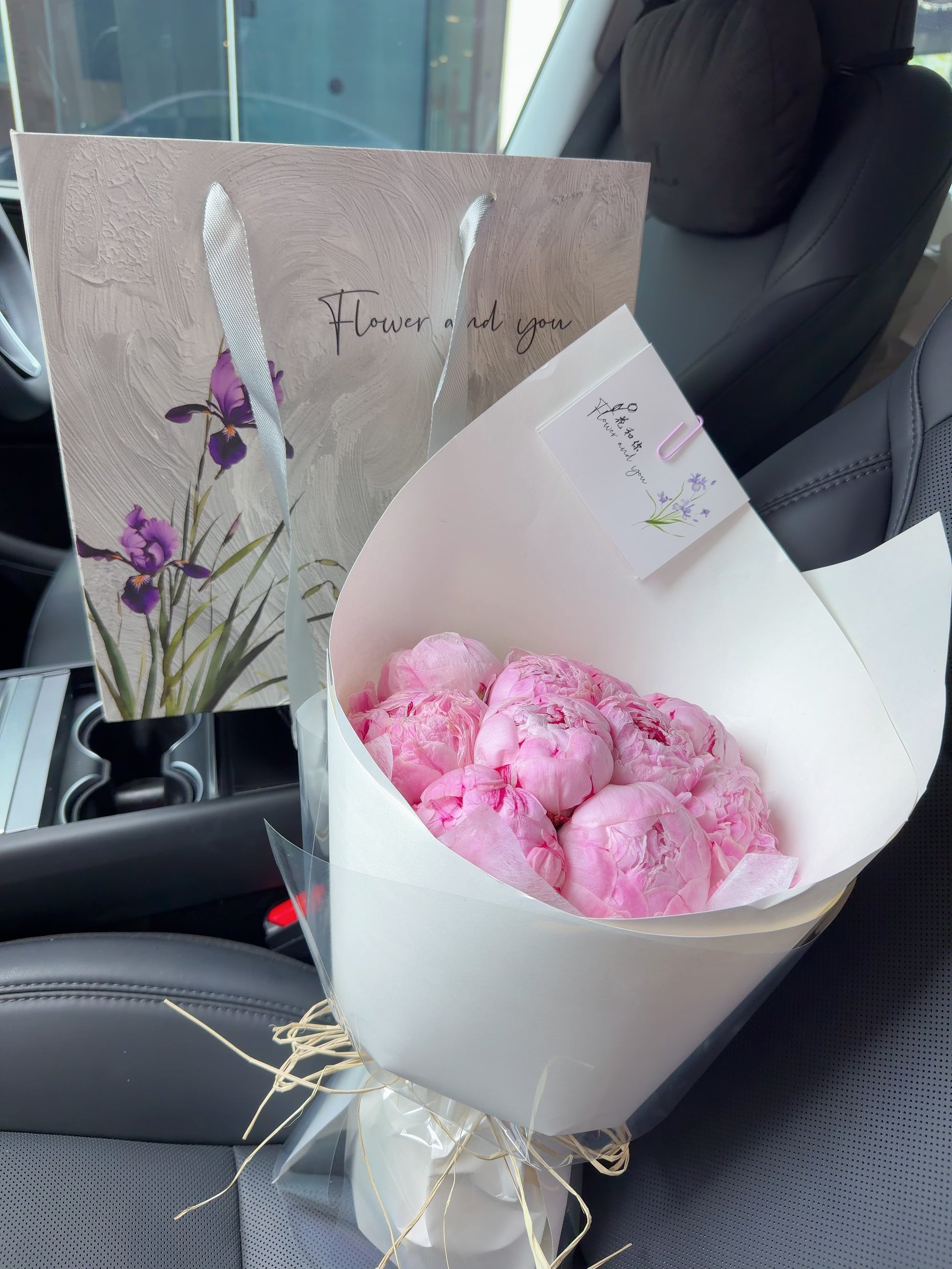 A bouquet of pink peony flowers wrapped in white paper sits on a car seat next to a printed gift bag with purple flowers and the words 'Flower and you'.