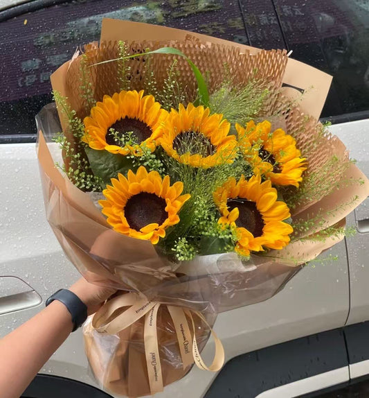 A bouquet of bright yellow sunflowers wrapped in brown paper and clear cellophane, held by a person's hand in front of a white car.