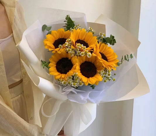 A bouquet of five yellow sunflowers with dark brown centers, accented with small white flowers and green leaves, wrapped in white and beige tissue paper and held by a person wearing a cream-colored top.