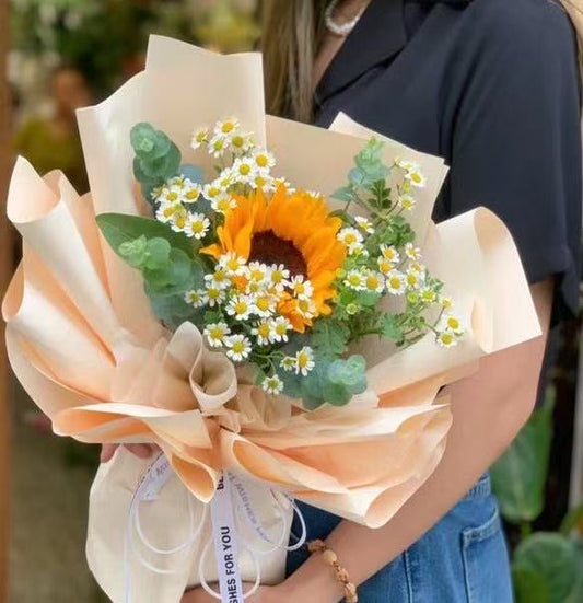 A person holding a wrapped flower bouquet featuring a large yellow sunflower, small white daisies, green eucalyptus, and green leaves, with peach and beige tissue paper wrapping and a white ribbon.