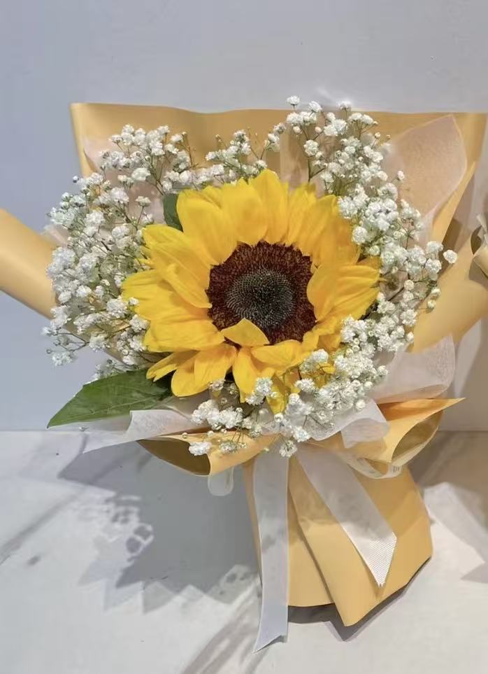 A single sunflower bouquet wrapped in pale yellow paper with white baby's breath and a white ribbon, photographed against a light neutral background.