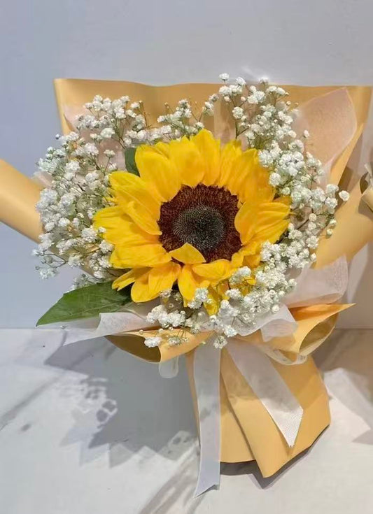 A single sunflower bouquet wrapped in pale yellow paper with white baby's breath and a white ribbon, photographed against a light neutral background.
