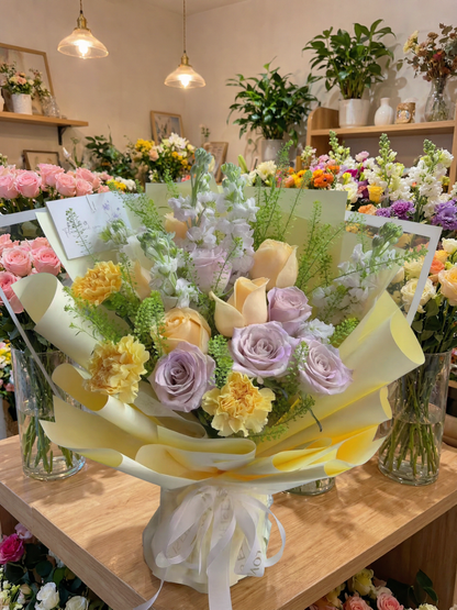 A pastel flower bouquet wrapped in pale yellow paper and tied with a white ribbon, displayed on a wooden table inside a flower shop. The bouquet features lavender roses, pale peach roses, yellow carnations, and green foliage.