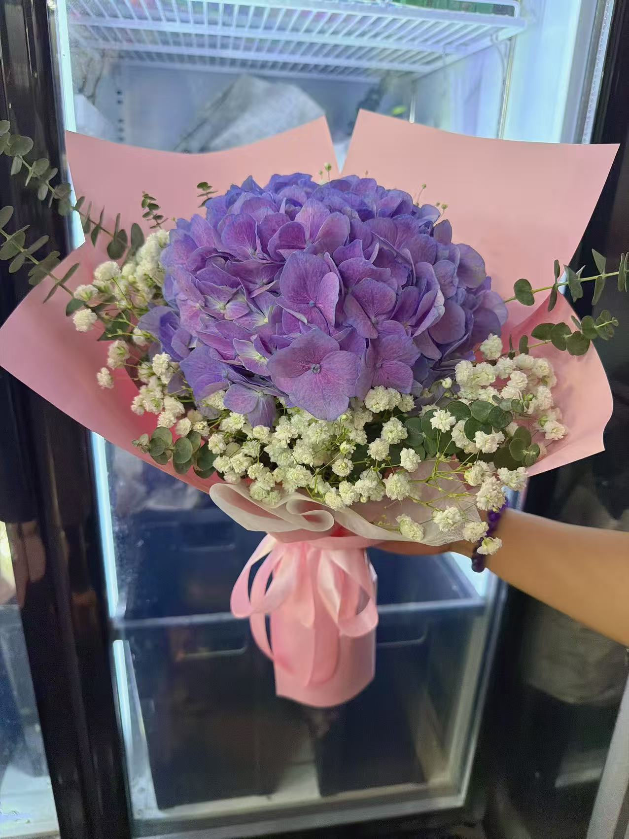 A bouquet of purple hydrangea flowers with white baby's breath and green eucalyptus stems, wrapped in pink paper and held by a hand in front of a glass display case.